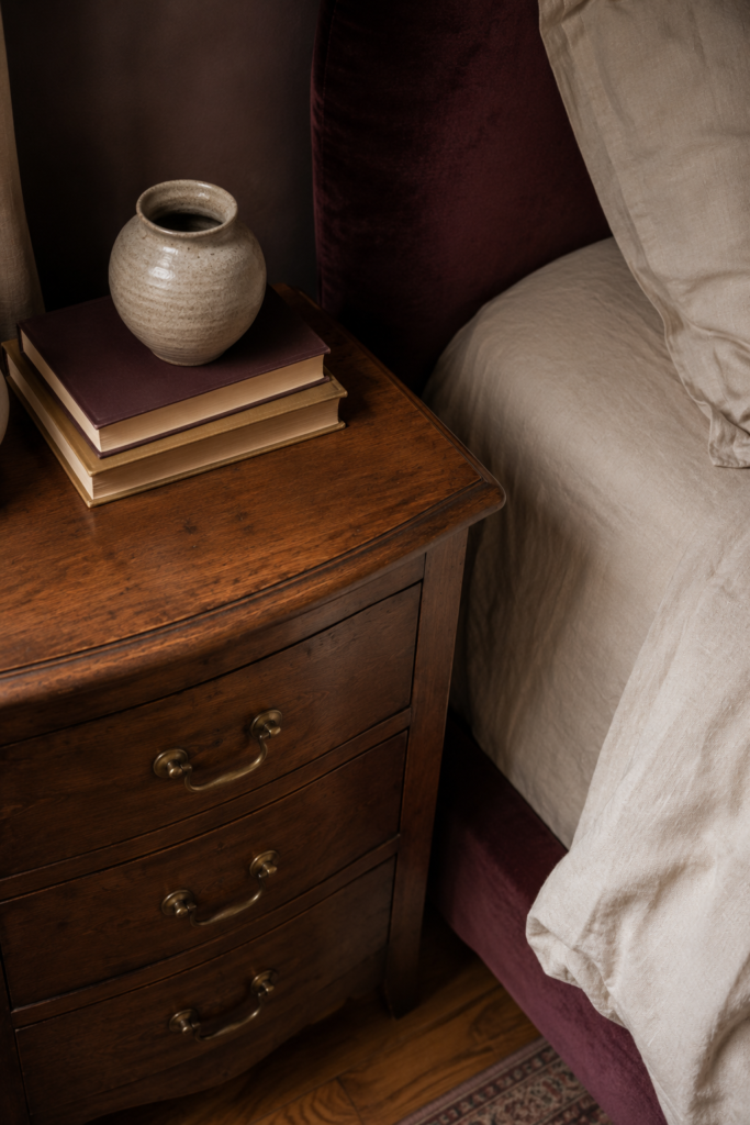 Vintage-inspired wooden nightstand with brass hardware beside relaxed cream bedding in a dark feminine bedroom.